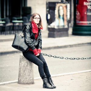 Danielle Bouchard, a woman with glasses, wearing a black leather jacket, red scarf, black pants, and black boots, sits on a concrete bollard on a city sidewalk. She has a large black handbag and is looking directly at the camera. The background shows out-of-focus urban elements.