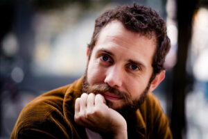 Grégoire Brossard, a man with short curly hair and a beard, rests his chin on his hand, looking directly at the camera with a slight smile. He is wearing a brown jacket and is outdoors with a blurred background.
