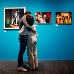 Raphael and Sophie embrace in front of a blue wall displaying three framed photographs in an art gallery. Sophie, in white pants and a sweater, stands on tiptoe, holding Raphael, who's dressed in jeans and a gray jacket. Two of the photos show scenes with people, and the third is abstract.