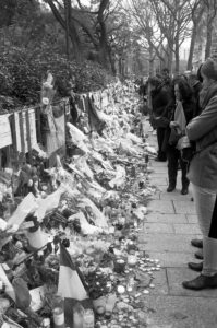 Black and white photo of a memorial site on a sidewalk. People stand in a line, observing flowers, candles, messages, and flags placed in tribute. The background features trees and more onlookers paying their respects.