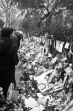 A black and white image shows people standing in silence, paying their respects at a memorial site. The memorial is adorned with numerous flowers, candles, and handwritten notes, creating a poignant scene of collective mourning and tribute.