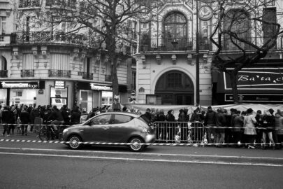 Black-and-white photo depicting a long queue of people standing behind barriers on a sidewalk, waiting outside historic buildings with ornate facades. A small car is parked in front of the crowd. The scene appears to be in an urban area, possibly a city center.