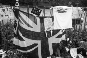 A black and white photo of a memorial site featuring a British flag, a white sports jersey with "Amlin" on it, handwritten notes, flowers, and various banners attached to a fence. The area is surrounded by greenery and a visible bench in the background.