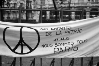 A banner is tied to a metal fence. It displays a peace symbol and French text that reads: "Aux enfants de la patrie 13.11.15 Nous sommes tous Paris," meaning "To the children of the nation 13.11.15 We are all Paris." The background shows a building.