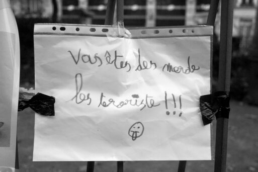 A handwritten note taped to a fence reads in French: "Vous êtes des merde les terroriste!!!" accompanied by a small drawing of a sad face. The note is held in a plastic sleeve and affixed with black tape. The surroundings are blurred.