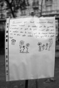 A handwritten note in French is displayed on a fence, enclosed in a plastic sleeve. The note is accompanied by drawings of flowers and a small, seated person. In the background, there are trees and a building, suggesting a park or outdoor setting.