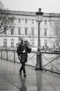 Black and white photo of Clara Hoareau walking on a wet bridge in the rain, holding an umbrella. She is dressed in a dark coat and pants. In the background, a tall historical building with numerous windows stands majestically. The bridge features an ornate lamppost and metal railings.