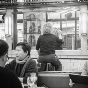 A person with curly hair, wearing dark clothing, stands on a chair facing a mirror in a café, writing on it with chalk. Between two chairs, two other people sit at a table in the foreground, engaged in conversation. The café is decorated with tiled walls and a "non fumeur" sign is visible.