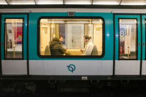 A nighttime view of a subway car, with two passengers seated and engaged in reading their lecture matinale. The car's windows reveal a bright interior contrasting the dark exterior. The subway walls and advertisements are visible through the windows.