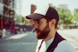 A man with a beard, resembling Grégoire BROSSARD, is wearing a black and white trucker hat and a white shirt while standing outdoors. He has a camera strap over his shoulder and is looking down. The background is blurred, showing buildings, trees, and scaffolding.