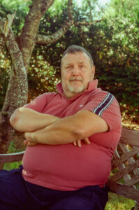 Marc LEPAGE, a man with short hair and a beard, sits on a wooden bench outdoors. He is wearing a red polo shirt with navy blue striped sleeves and has his arms crossed. The bench is placed under a tree, with green foliage visible in the background. The sun appears to be shining.