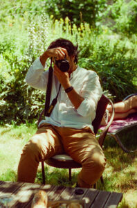 Daniel, in a white shirt and brown pants, is sitting on a chair outdoors, holding a camera up to his face, taking a photo. The background features lush green foliage. Another individual, LATIF, is lounging on a chair nearby, partially visible.