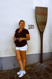 A woman in a white shirt and shorts stands against a white wall with a plaque written in French next to her. She holds a camera around her neck and smiles at the camera. The wall, marked with "SYSTEME LAVRAC," resembles the artistic touches seen in Emmanuelle RADENAC's work.
