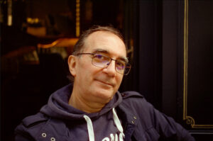 A man with glasses and graying hair, wearing a navy jacket and a hoodie, is looking at the camera with a slight smile. The background appears to be a dark indoor setting with soft, warm lighting, creating an atmosphere reminiscent of Helder VINAGRE’s artistic sensibilities.