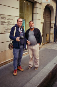 Two men are standing outside a building on a city street. The man on the left, Pascal HAWLIK, is wearing glasses, a blue vest, blue jeans, and tan boots, with a camera around his neck. The man on the right, Helder VINAGRE, is wearing a dark jacket, a gray shirt, and beige pants. Both are smiling.