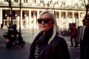 Annalise Nogena, a woman with long blonde hair wearing sunglasses and a dark coat, stands outdoors on a sunny day. There is a blurred background of a city street with people walking and a person on a motorcycle.