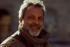 A man with gray hair and a beard smiles warmly at the camera. He is wearing a light brown jacket and a scarf. The background is blurred, suggesting an outdoor setting, possibly in an urban area. This charming scene captures Pascal Hawlik in his natural element, radiating warmth and friendliness.
