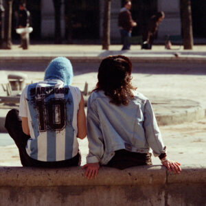 Two people sit on a low concrete wall during the day. One, with a blue hood and light blue and white striped shirt displaying "Numero 10," represents Argentina's iconic colors. The other person, with dark hair, is sporting a light denim jacket. People are seen walking in the background.
