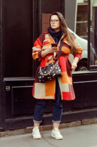 Nadia Le Roux, with her long hair flowing, stands outdoors in front of a dark wall, holding a smartphone. Sporting glasses and a red and orange checkered coat, she pairs rolled-up jeans with white sneakers. Her black purse with a chain strap and white polka dots adds the final touch to her chic look.