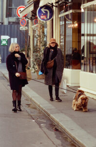 Two women are walking down a city street. One woman, wearing a black coat and boots, is looking at her phone. The other woman, dressed in a long coat and cap, is walking her brown dog, named Chien Luka, on a leash. Storefronts and street signs are visible in the background.