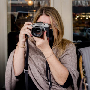 A woman photographer with long blonde hair is aiming a silver Fujifilm camera at the viewer. She is seated at a table, adorned in a shawl and jewelry, with indoor lighting suggesting the cozy ambiance of a café or restaurant.