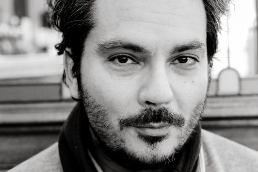 20240302-DSCF1500_MOD_BW_FRAME_resize Black-and-white close-up portrait of Daniel Latif, a man with short, dark, and slightly graying hair. He has a beard and mustache and is looking directly at the camera with a neutral expression. He is wearing a scarf and a casual jacket. The background is blurred.