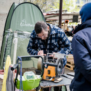 An older man in a checkered jacket, possibly Robert Mounier, focuses on repairing a sewing machine at an outdoor market. In the background, a green tent with the logo "Wolfwise" is visible. Various market stands and people can be seen bustling around.