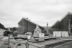A large construction site with extensive scaffolding and several temporary structures. A historical monument is partially visible surrounded by construction materials and fenced-off areas. The sky is overcast, and there are signs indicating safety instructions.