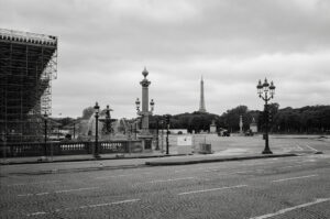 Black and white image of Place de la Concorde in Paris, featuring fountains, ornate street lamps, and scaffolding on a building in the foreground. The Eiffel Tower is visible in the distance under a cloudy sky. The square is notably empty.