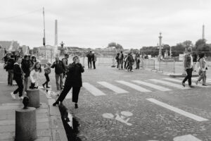 A black and white photo of people walking and standing near a crosswalk on a wide cobblestone street. Some are taking photos, while others are casually strolling. Various architectural landmarks and a distant tower are visible under a cloudy sky.