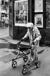 An elderly person using a walker navigates a city sidewalk near a bus stop or advertisement stand. In the background, there is a large poster featuring French athlete Teddy Riner, dressed in white. The cityscape of France subtly adds to the scene's charm and vibrancy.