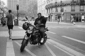 Ivan Bonneau is sitting on a motorcycle parked on the side of a city street, adjusting his helmet. Several pedestrians are visible in the background, near buildings and a crosswalk. The scene is in grayscale, portraying an urban environment.