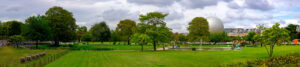 A panoramic view of the large, lush Jardin des Tuileries with green lawns, various trees, and a vasque olympique fountain in the center. In the background, buildings and a large spherical structure are visible under a cloudy sky. A few people can be seen walking and enjoying the park.