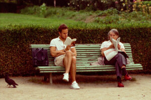A man and an older woman sit on a park bench, each absorbed in reading a book. A black bag rests beside the man, and a pigeon stands on the ground nearby. The background features a hedge and a grassy area.