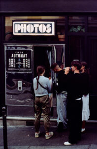 Four people stand in front of a vintage photo booth labeled "PHOTOS" on a dark street, reminiscent of an art piece titled Photo Booth. A sign above lists pricing and instructions. They prepare to take pictures, with one person clutching a camera.