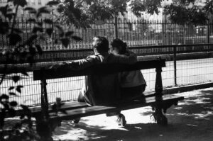 A black and white photo of Justin and Georgette sitting closely on a park bench, with Justin resting his arm on the back. They face a fence and trees, as sunlight casts shadows on the ground.