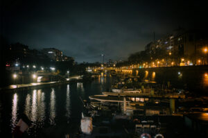 The Port de l'Arsenal presents a tranquil nighttime scene with boats docked along the city canal. Streetlights shimmer on the water's surface, while buildings stand solemnly under the dark, overcast sky of this serene nuit.