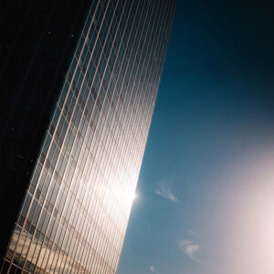 A tall glass skyscraper in La Défense reflects sunlight, creating a bright glare on its surface. The sky is clear with a deep blue gradient. The sun is partially visible near the lower right corner, casting soft light across the scene, reminiscent of Tour Initiale’s iconic presence.
