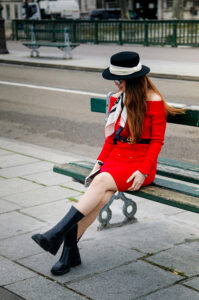 A woman in a robe rouge and black boots sits on a bench. She wears a wide-brimmed chapeau noir and a scarf, with long hair cascading down. The street and a green railing are visible in the background.