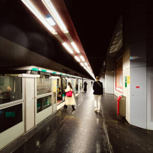 A dimly lit subway station with a train stopped at the platform. Two people walk along the platform: one in a long beige coat, the other in a black jacket and white pants. The station has a modern design with overhead lighting.
