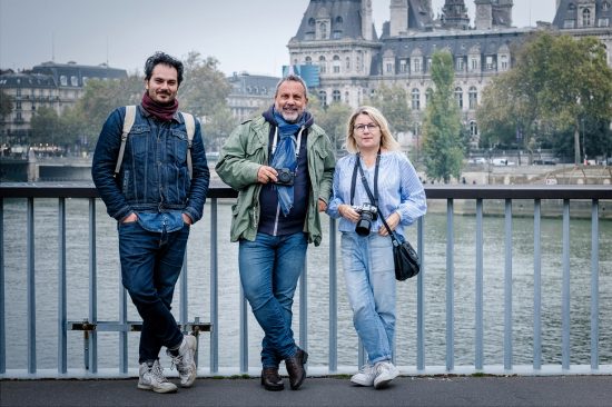 Three people with cameras stand on a bridge, smiling in a casual, relaxed manner. Behind them is a river and a large historic building. The sky is overcast, creating a cool atmosphere. They're dressed for cooler weather.