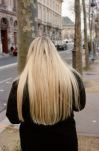 Florence Schwartz, with her long blonde hair, strolls down a city sidewalk lined with trees and historic buildings. The street is quiet, the sky overcast, drawing attention to the texture and length of her hair.