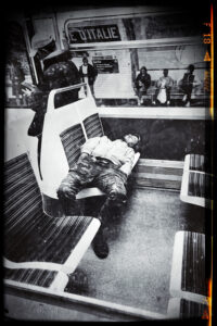 A man is sprawled across two seats inside a subway train on Ligne 7, seemingly asleep. Outside the window, passengers wait at Place d'Italie station. The black-and-white image, with a vintage filter, captures a timeless moment of city life.