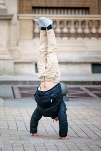 Vladimir Hamelin, in a black jacket and beige pants, performs a handstand on a paved outdoor area with a stone building in the background.