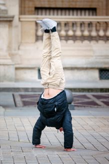 Vladimir Hamelin, in a black jacket and beige pants, performs a handstand on a paved outdoor area with a stone building in the background.