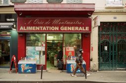 A man walks past a small grocery store labeled "Alimentation Generale" with a red facade. The entrance displays a variety of products inside and ice cream freezers outside. The store is situated between a green-paneled door and a shop offering tacos and paninis.