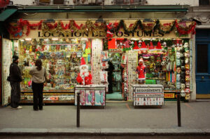 A colorful costume shop with a festive display, featuring Christmas decorations, masks, and various costumes. Two people stand outside browsing. Signs in French advertise costume rentals and sales.