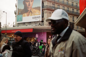 Street scene with a crowd of people walking past a busy intersection. A large Versace advertisement featuring a model is visible on a building above. Some are wearing winter clothing, and bicycles are parked nearby. Urban setting with overcast weather.