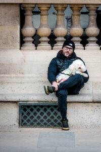 A person wearing a black beanie, glasses, and a black jacket sits on a stone ledge with Edgard Leclercq's signature style. They hold a small white dog in a colorful outfit, framed by classic stone balusters in the background.