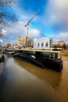 Construction crane near Notre-Dame Cathedral in Paris, on a cloudy day. A barge is docked along the Seine River, and a large billboard hangs on the cathedral's facade. Bare trees line the riverbank.